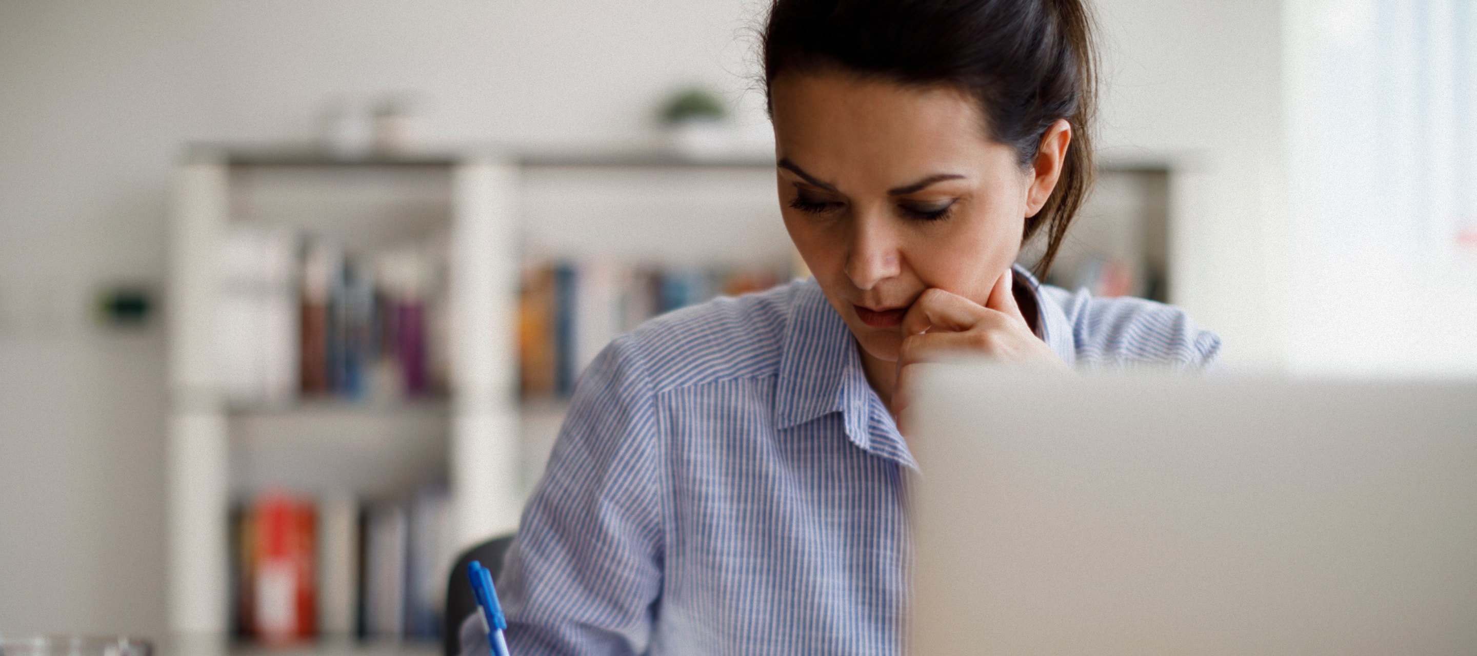 Woman looking down with blurry laptop in foreground and books in background.